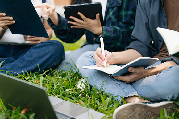 Auf einer Wiese sitzen verschiedene Menschen mit Büchern und Tablets, die Gesichter sind nicht zu sehen, weil das Bild an den Schultern endet